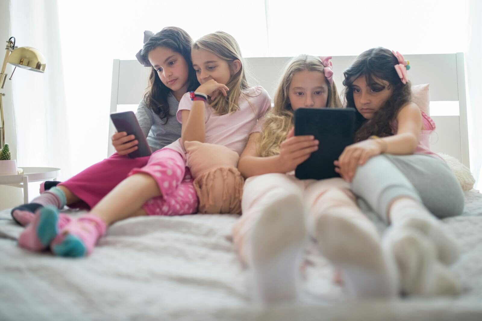 Four girls sitting on a bed using tablets, engaged and relaxed in a cozy indoor environment.